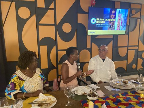 Two Black women and one Black man sitting at a wooden dining table. The Black woman in the middle is talking into a microphone.
