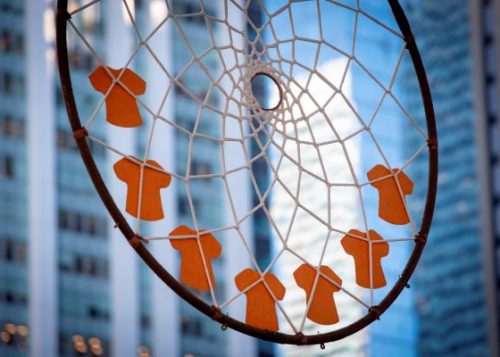 A dreamcatcher showing orange shirts in commemoration of Indigenous truth and reconciliation with city buildings in background.