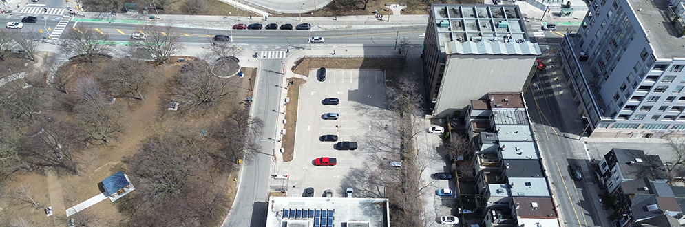 An aerial view showing a parking lot and the building directly south of it, designated as the future site of a Community Recreation Centre. To the upper right, the southeast corner of Ramsden Park is visible, including picnic areas surrounded by trees and pathways. The image also captures Rosedale TTC station and its loop, along with nearby streets, commercial spaces, and low-rise residential buildings.