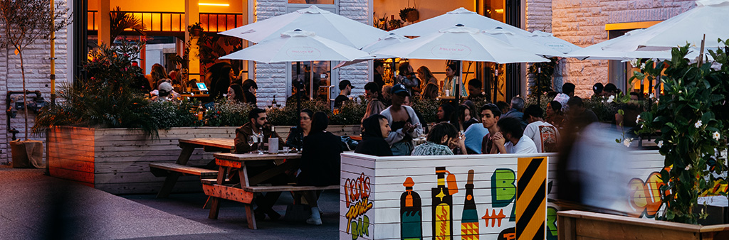 A busy restaurant patio in the evening with picnic tables, white umbrellas and colourfully illustrated and painted fencing.