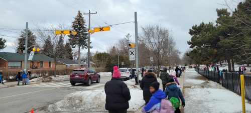 A local road with a pedestrian crosswalk and people including school children walking on the sidewalk and people waiting to cross the crosswalk as a car drives by.