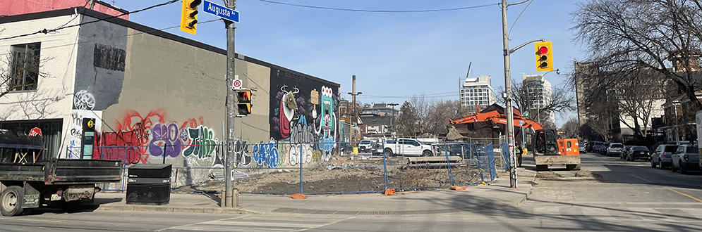 Photo of the future park showing a fenced in empty lot at the corner of Queen Street West and Augusta Avenue, next to a building with graffiti along the wall.