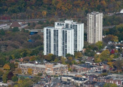 Older high-rise apartment buildings overlooking houses and other buildings with trees and a highway in the background.