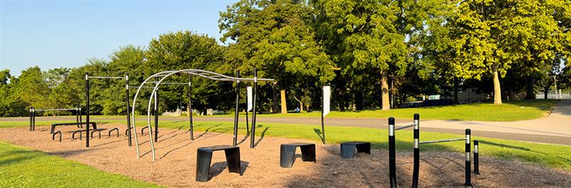 An outdoor fitness area with workout equipment, surrounded by lush green trees and a paved pathway.