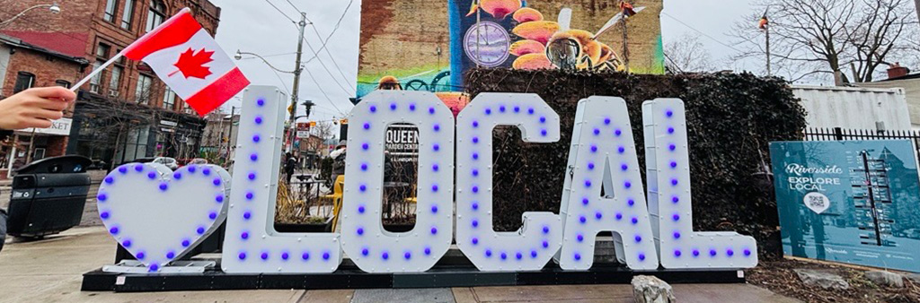 A marquee letter sign, white with blue lights that starts with a heart and is followed by LOCAL. A canadian flag is held out in front of the sign which sits in a public parkette.