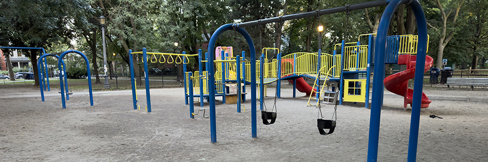 A playground in a park featuring blue and yellow play structures, including swings, monkey bars, and slides. The ground is covered in compacted dirt, with visible pathways worn by foot traffic. Trees surround the area, and a fence encloses part of the playground. In the background, there are park benches and lampposts.