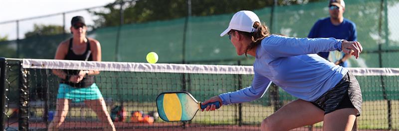 A group of players engaged in a lively pickleball match on an outdoor court, with a player in a white cap and blue long-sleeve shirt reaching to hit the ball with a paddle.