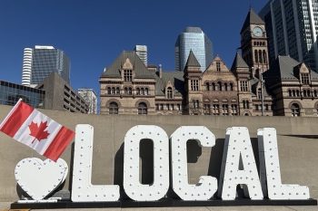 A white marquee letter sign that starts with a heart and is followed by LOCAL. A canadian flag is held out in front of the sign which sits in the public square with a view Old City Hall in Toronto in the backgound.