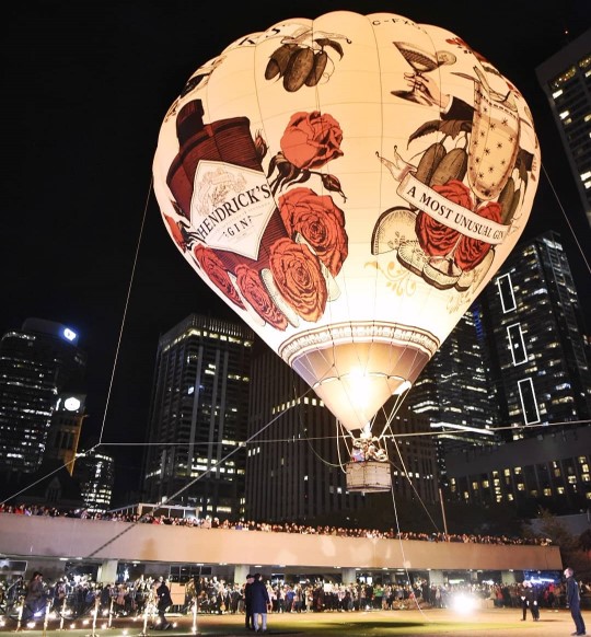 A large, illuminated hot air balloon featuring Hendrick’s Gin branding and artwork floats at night with background of skyscrapers. Crowds of spectators gather below and on the surrounding ramp.