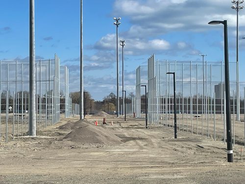 View of the Centennial Park baseball hub under construction. The photo shows rows of tall metal fencing frames for baseball diamonds, newly installed light posts, and mounds of gravel across a dirt-covered area. The scene is set under a blue sky with scattered clouds, and some traffic cones are visible marking construction areas.