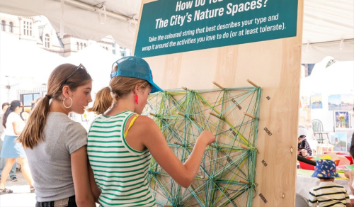 Two people interact with a peg board with string as part of an interactive installation titled how do you use the City's Nature Spaces?