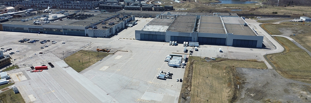 Aerial view of the Downsview Airport lands in Toronto, featuring large industrial hangars and warehouse-style buildings with numbered doors (1 to 5) along a wide expanse of paved tarmac. The area is surrounded by open fields, patches of grass, and construction equipment. Several transport trucks and trailers are parked near the buildings. In the background, mid-rise residential buildings and a pond are visible, along with a broader view of the city skyline in the distance. The photo captures the scale and industrial nature of the site amidst ongoing redevelopment.