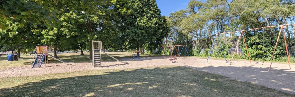 A playground in a grassy park setting with mature trees in the background. The playground features a slide structure on the left, a smaller climbing structure in the center, and two swing sets on the right, all set in a large sand play area. The area is surrounded by open lawn and shaded by large leafy trees.