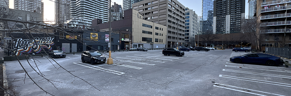 A nearly empty urban parking lot surrounded by high-rise buildings in the background. A few cars are parked in various spots, and the pavement has visible cracks and patches of ice or snow. A mural with the words "Yonge Street" in colorful graffiti is painted on a black building to the left. Leafless tree branches extend into the frame from the foreground. The sky is clear, with a soft glow from the setting or rising sun reflecting off the glass skyscrapers.