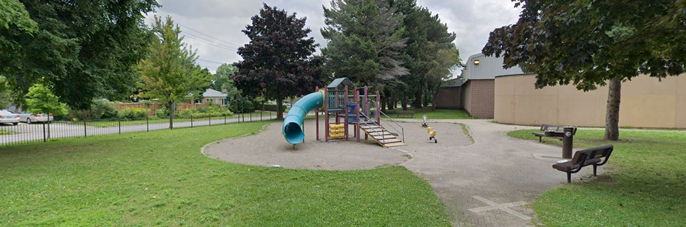 A small playground set in a grassy park space beside a community building. The play structure includes stairs, platforms, and a blue spiral slide, with two spring rider toys nearby. The area is surrounded by mature trees, a fence along the street, and paved walkways with benches on either side.