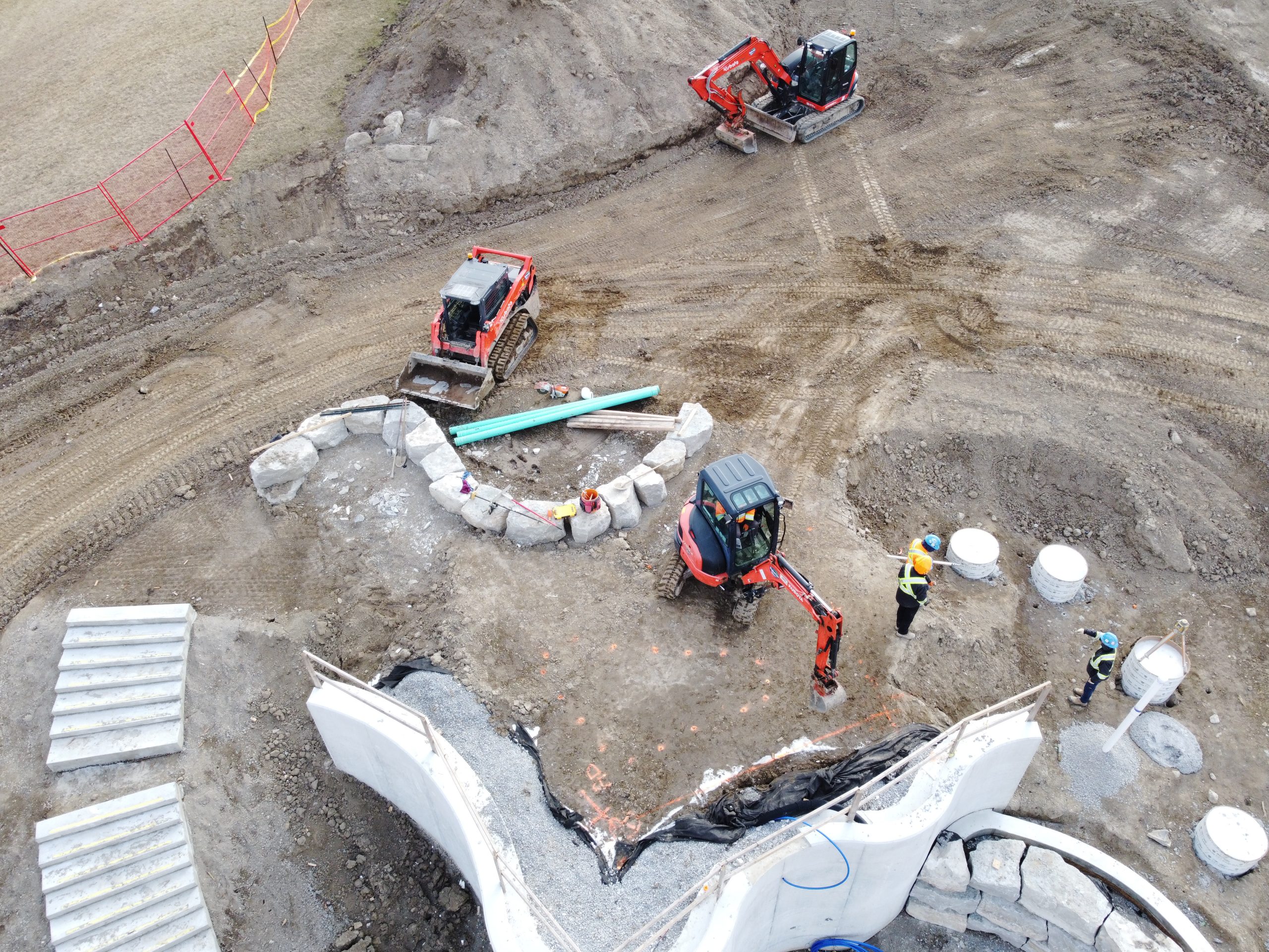 An aerial close-up of the waterfall area in the water park. From left to right are steps leading to the top of the waterfall and footings for the hill water slide, both made of concrete. Three excavators are parked in the area, including one positioned to dig near a shallow pit where construction workers stand in safety gear. Piles of gravel and construction materials are scattered on the dirt ground. Some orange fencing is visible along the upper-left edge of the site, separating it from an open grassy area.