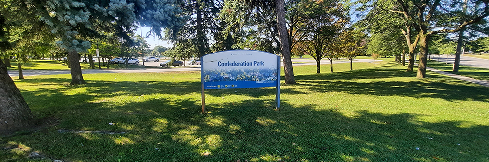 The Confederation Park identification sign on top of a green open lawn areas with mature trees and the sun shining in the background.