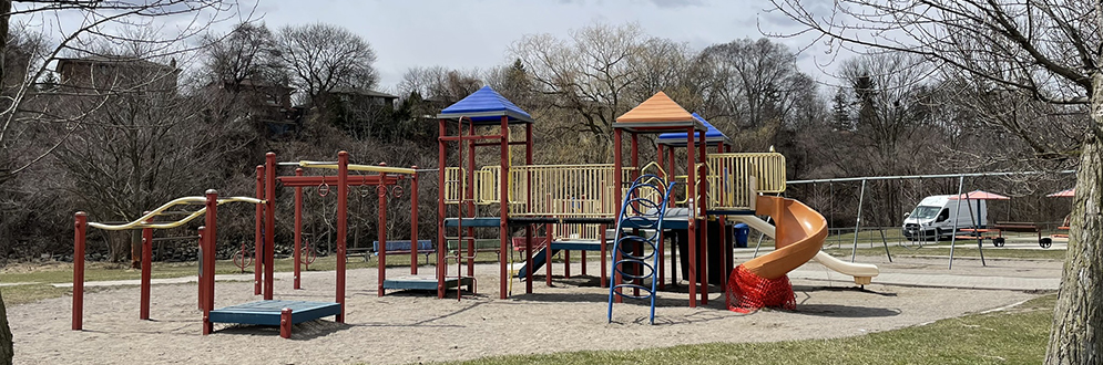 Weston Lions Park Playground showing a large play structure in red, blue and yellow with monkey bars, climbing features and two slides. A swing set is in the background. The playground is on top of sand.