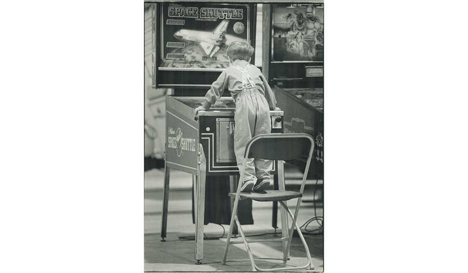 Child stands on a folding chair to reach and play an arcade game