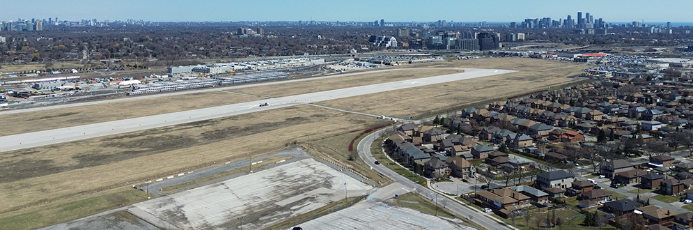 Aerial view of the Downsview Airport lands in Toronto, featuring a long runway running diagonally across the image. The runway is surrounded by large open grassy areas and bordered by industrial buildings, train yards, and low-rise suburban housing in the foreground. Mid-rise developments and construction sites are visible beyond the runway. In the distance, the Toronto skyline is clearly visible under a bright blue sky, with clusters of high-rise buildings marking the downtown core. The image captures a mix of industrial, residential, and future redevelopment areas.