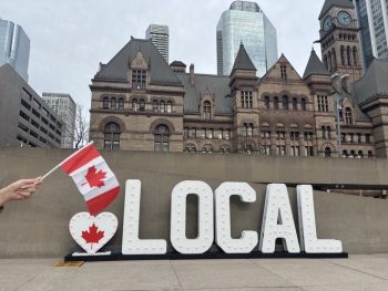 A white marquee letter sign that starts with a heart with a red maple leaf inside of it, and is followed by the letters LOCAL. A canadian flag is held out in front of the sign which sits in the public square with a view Old City Hall in Toronto in the background.