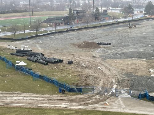 Aerial view of the soccer hub construction site at Centennial Park during light snowfall. The image shows a large dirt field with piles of gravel, construction materials such as stacked pipes, and a bulldozer. Blue construction fencing lines the perimeter, and a running track and building are visible in the background.