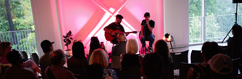 A singer and guitar player perform for an audience in front of a neon pink wall.
