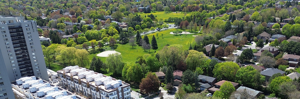 Aerial view of a suburban neighborhood bordered by a park. The image shows a mix of housing types and there is a winding road and many trees.