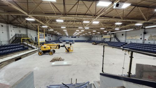 : A wide-angle view of an indoor ice rink under construction with the ice surface removed. Construction equipment including several lifts and wooden pallets are scattered across the concrete rink floor. Blue stands and brick walls surround the rink on all sides. Overhead lighting attached to exposed ceiling beams illuminates the space.