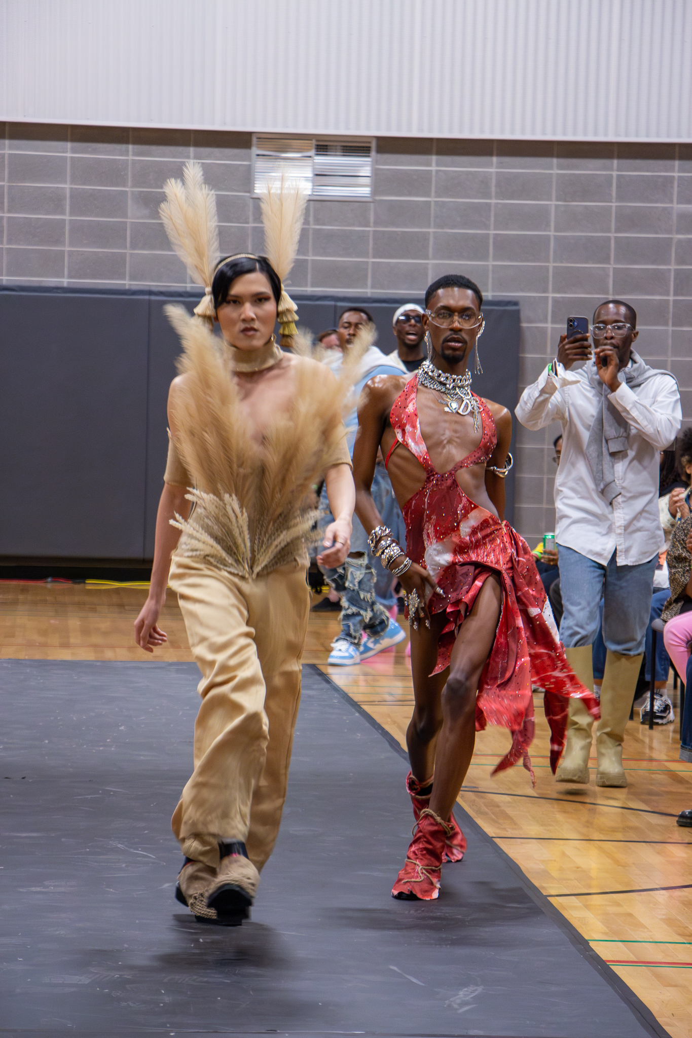 Two models walking on a runway during a fashion show. The model on the left wears beige pants and a top with large feather-like decorations. The model on the right wears a red, asymmetrical dress with cutouts and layered accessories. Spectators in the background are watching and taking photos