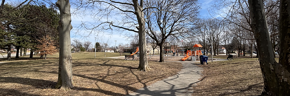 A paved pathway winds through a park with leafless trees casting long shadows on the ground. In the distance, an orange playground structure is visible, surrounded by benches and waste bins. The grass is brown, suggesting early spring or late fall. A few people are scattered throughout the park, enjoying the sunny but cool day.