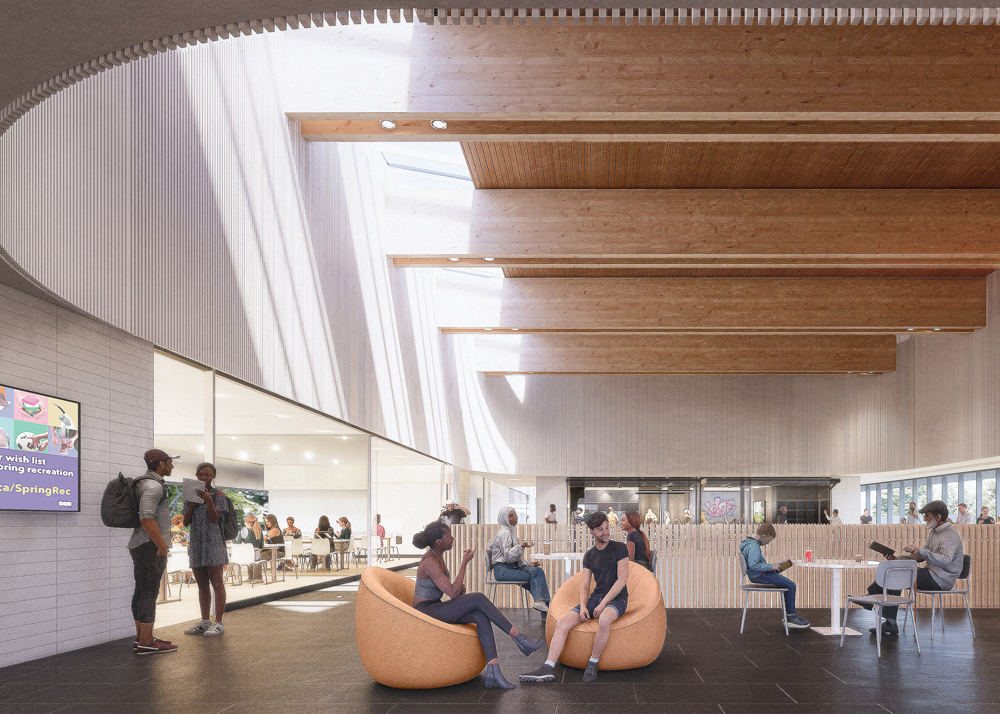 Rendering of the lounge in the community centre. The curved space has high ceilings with wooden beams and skylights on one side. People are shown sitting in round orange fabric chairs and in chairs around tables. A partial wooden fence provides separation in the centre of the room. Large glass windows are visible on three sides, providing views into adjacent rooms and trees outside.
