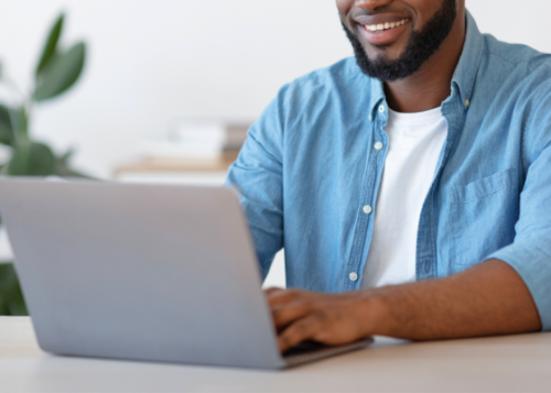 A person smiling as they type on a laptop with a plant in the background.