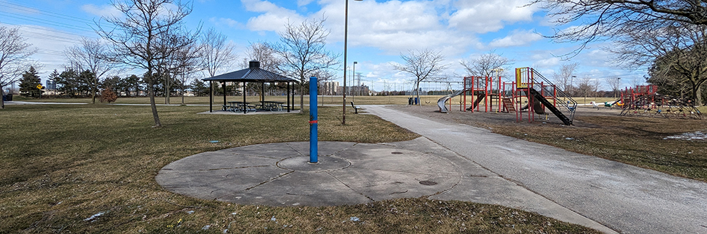 Jack Goodlad Park showing the existing splash pad and playground, looking northeast from the pathway. In the foreground is a small circular concrete pad with one blue spray feature. In the background is an open lawn with trees, a shade structure and various play structures.
