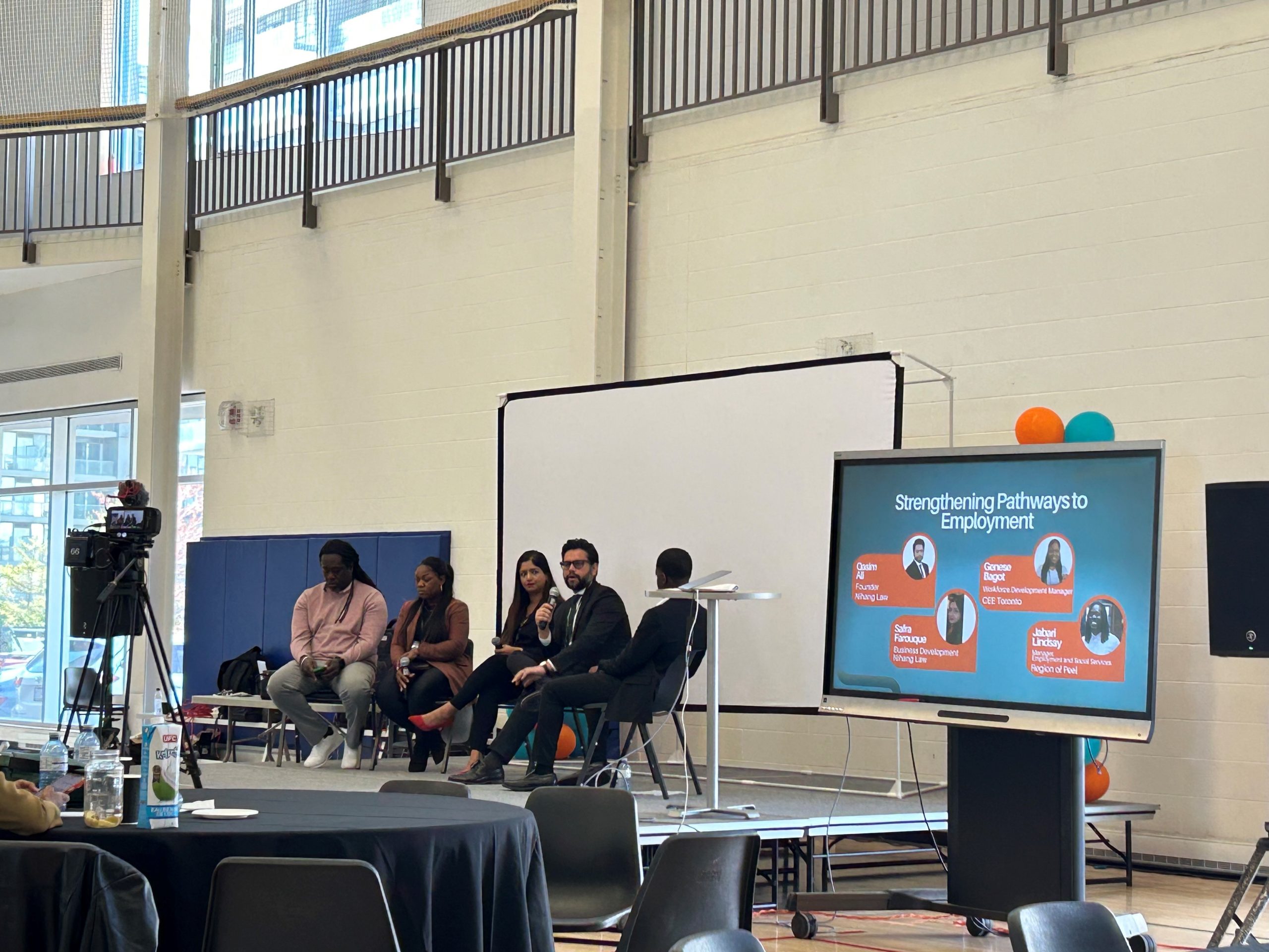 A panel of five panelists are engaged in a discussion. They are seated on a stage in a large room. There is a screen next to them displaying the title "Strengthening Pathways to Employment"