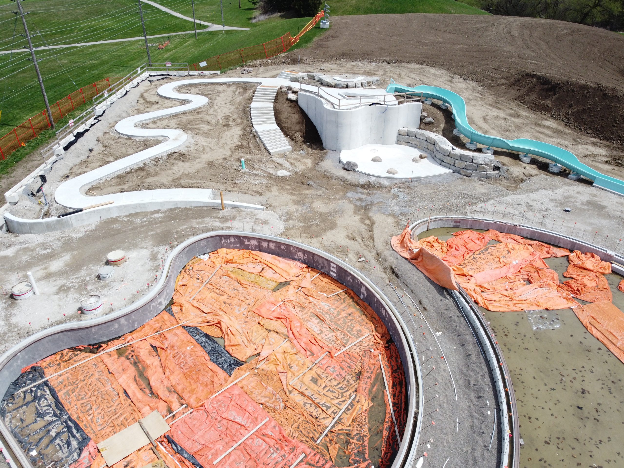 An aerial close-up of the pools and waterfall play area under construction, looking east. In the foreground, two curved pool basins are lined with concrete walls and covered with orange tarps. Past the pools, a winding ramp and set of steps, both made of concrete, lead to a curved structure forming the top of the waterfall area. A round concrete base and partial stone wall forms the base of the waterfall area. To the right, a wavy turquoise slide extends down on a slight slope. Orange fencing surrounds the construction site, and green grassy hills are visible in the background.