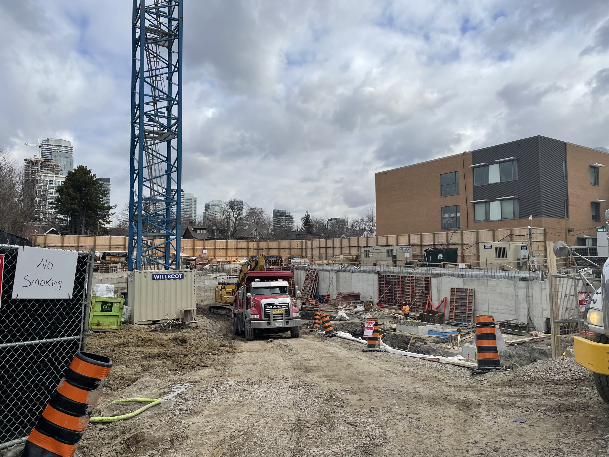 A fenced construction site with a partially built concrete foundation for a large building, adjacent to a multiple-storey brick building. A red truck and a blue crane tower are situated at the centre of the site, surrounded by various construction materials including safety bollards, storage containers and pallets. A worker wearing safety gear is visible by a foundation wall. In the foreground, a handwritten “No Smoking” sign is attached to a chain-link fence leading into the site. In the background, tall buildings and trees stand under a cloudy sky. 