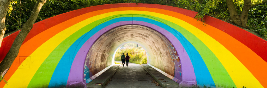 Two people in shadow on a walking trail through a tunnel, the outside of tunnel exit is painted as a large rainbow and is surrounded by greenery above.