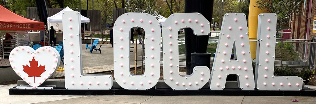A white block letters marquee sign that starts with a heart with a red maple leaf in the middle followed by the word LOCAL. The signs sits outside in an area with trees.