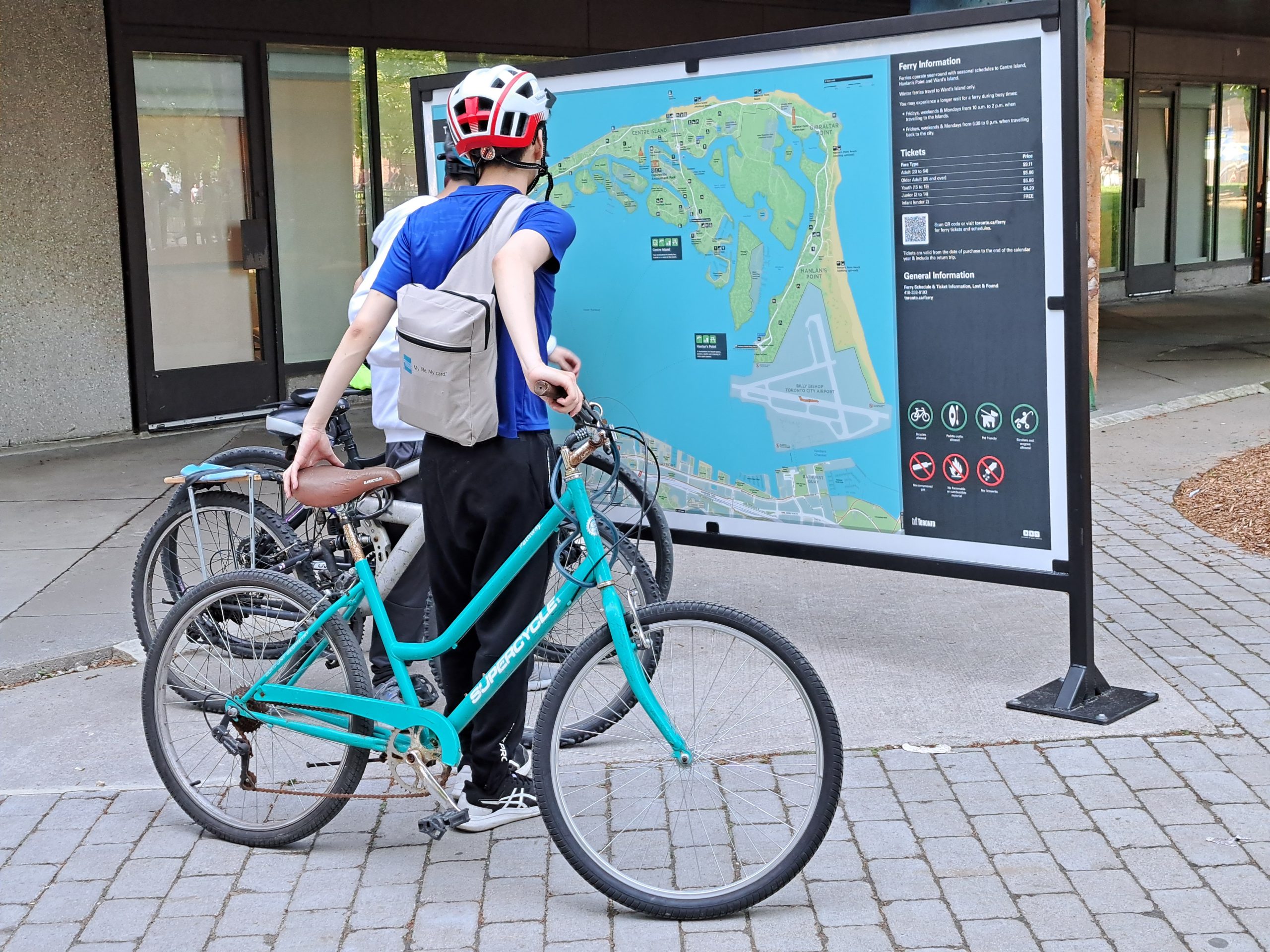Three passengers with bicycles look at the large map of Toronto Island Park located at the entrance to Jack Layton Ferry Terminal.
