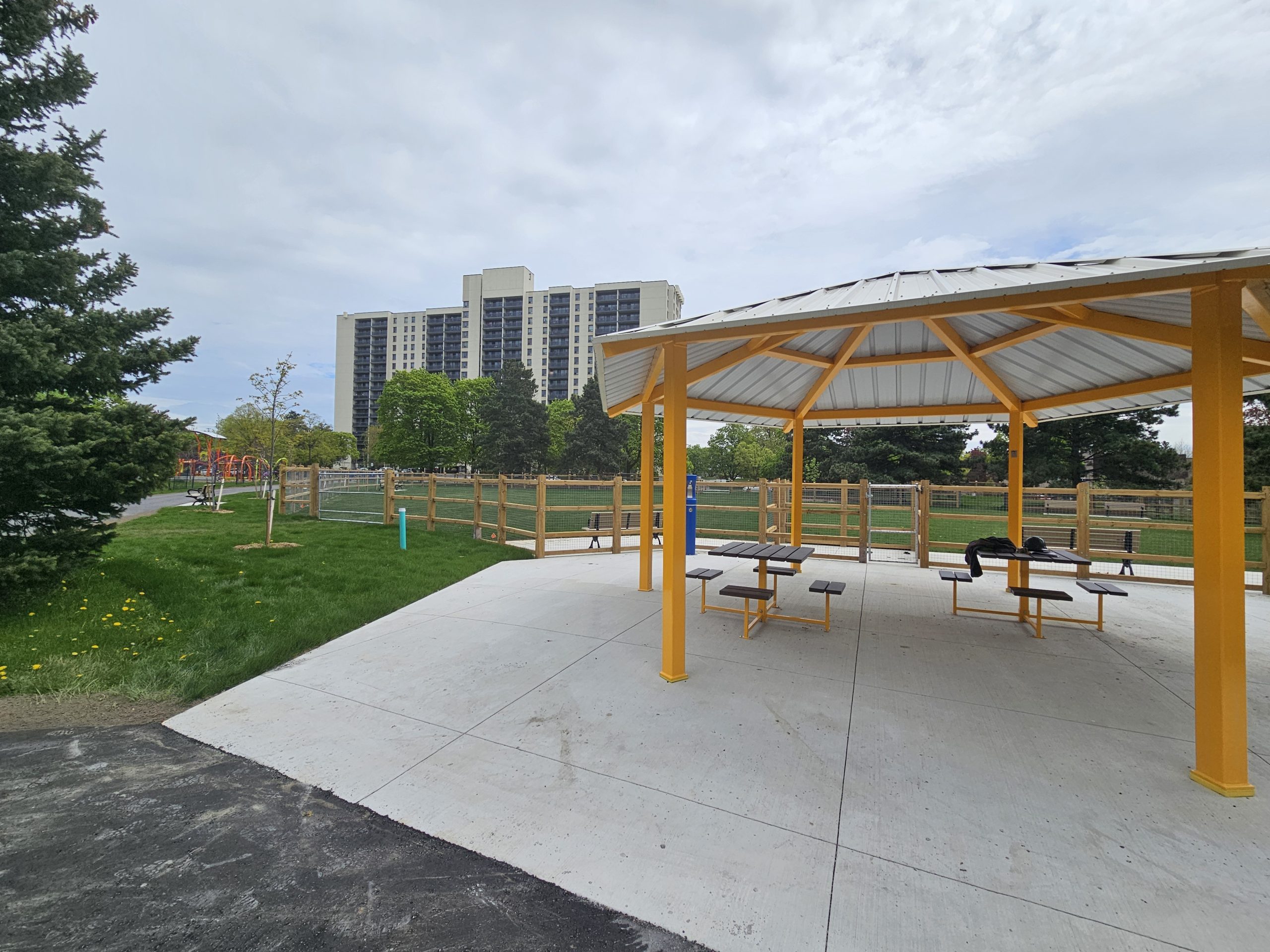 A view of the new off-leash area in progress, with the open-sided shade structure in the foreground. The structure’s grey roof is supported by yellow poles that stand on a concrete pad. Under the shade structure are two newly installed tables, each with four attached chairs. Behind the structure is an open grassy area surrounded by a wood and wire fence. A park playground and trees are visible in the background, along with residential buildings further in the distance.