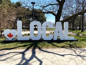 A white block letters marquee sign that starts with a heart with a red maple leaf in the middle followed by the word LOCAL. The signs sits outside in a park area with trees on a sunny day.