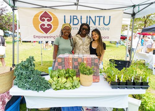 Three people standing behind a table selling plants and other production at an outdoor market. A sign for their business hangs behind them.