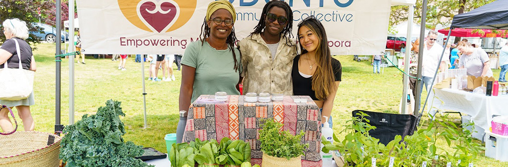 Three people standing behind a table selling plants and other production at an outdoor market. A sign for their business hangs behind them.