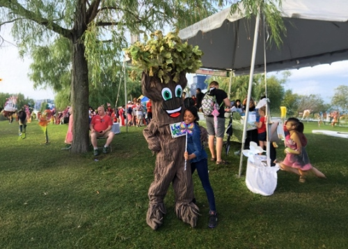 A child hugs a person dressed up as a tree and holds a Live Green Toronto sign with a lawn set up with tables, chairs and a tent in the background.