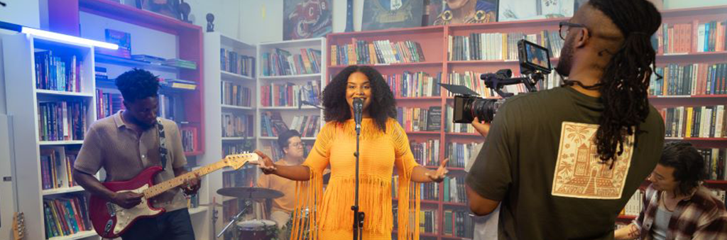 A woman in a yellow dress sings at a microphone with bookshelves behind her.