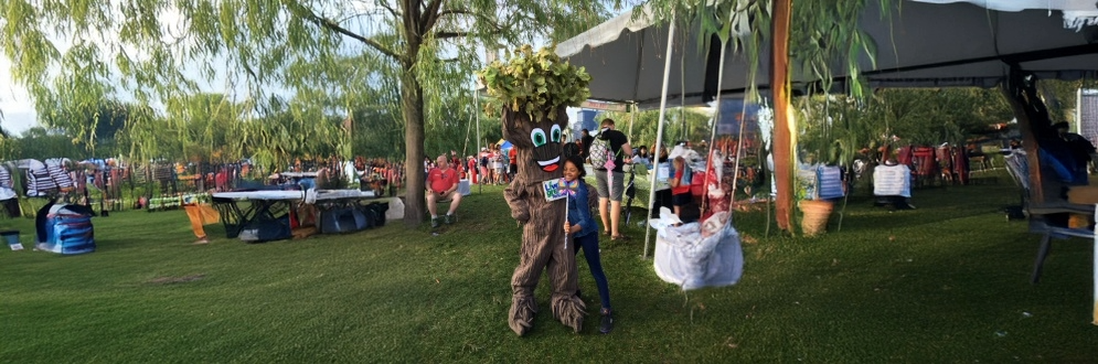 A child hugs a person dressed up as a tree and holds a Live Green Toronto sign with a lawn set up with tables, chairs and a tent in the background.