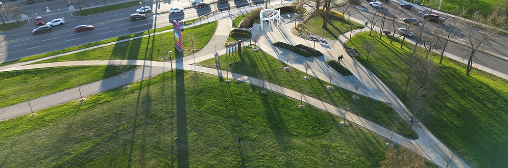 Aerial view of the park, highlighting the white pavilion, signage, and pathways. A tall pink and multicolour vertical sign is prominently visible near the street.