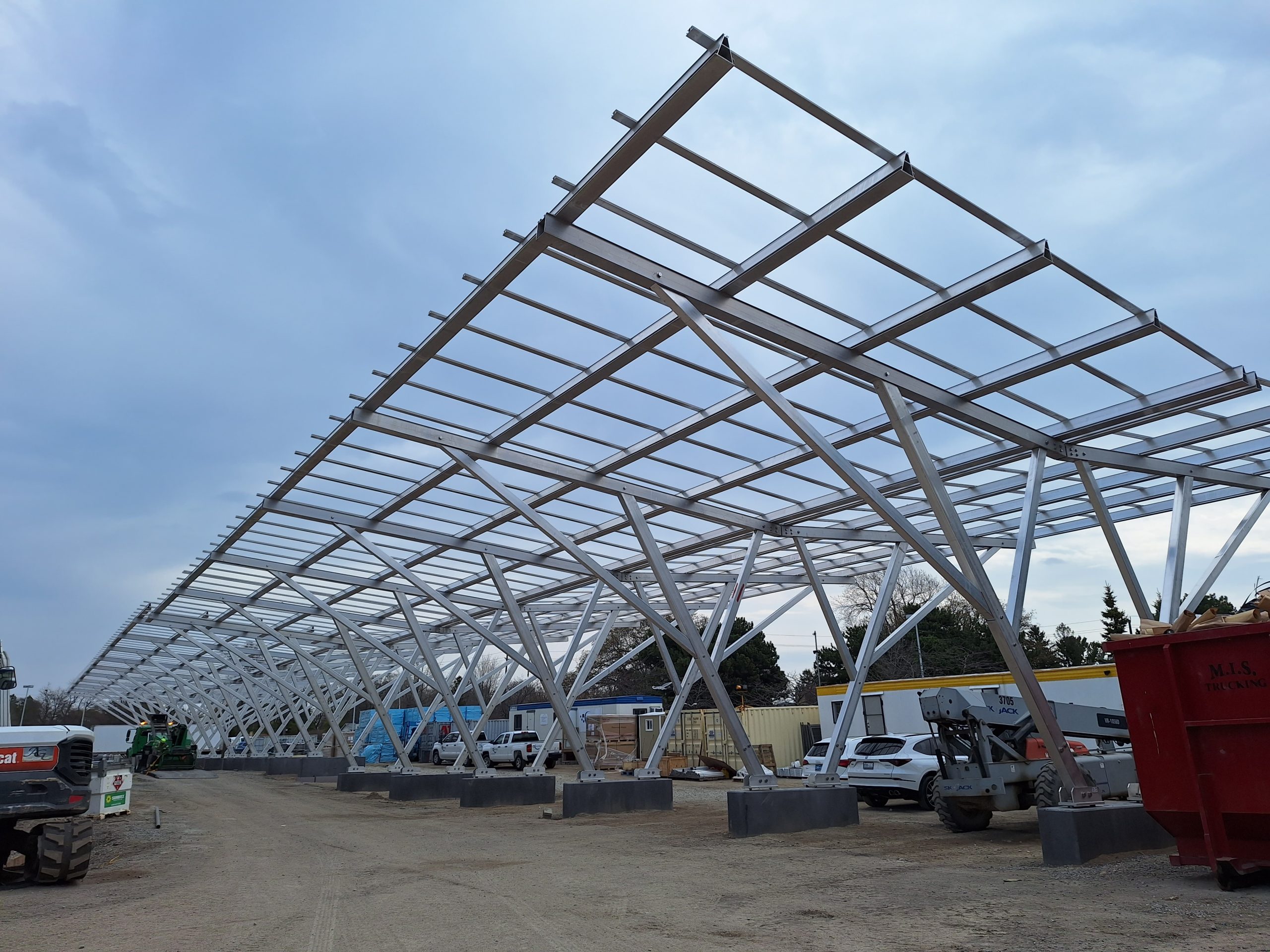 A ground-level photograph showing the solar carport structure in progress. The large metal framework is supported by angled beams attached to concrete bases and extends horizontally along a dirt construction site. Construction equipment, parked vehicles and scattered piles of construction material are visible around the