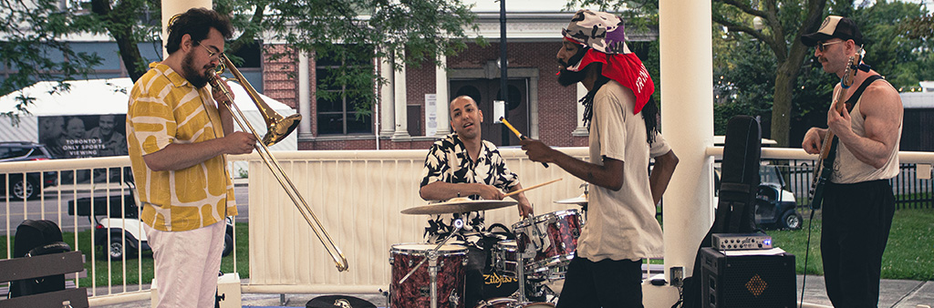 A band of four including a drummer, bass player, trombone player perform on a small outdoor stage.